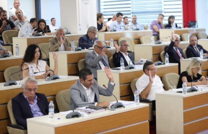 Görselde bir belediye meclis toplantısı görülüyor. Salon sıralı oturma düzeninde; önlerindeki mikrofon ve belgelerle meclis üyeleri toplantıya katılıyor. Birçok kişi ellerini kaldırarak oylamaya katılmış. Arka sıralarda izleyiciler de yer almakta. Resmi ve ciddi bir ortam dikkat çekiyor.