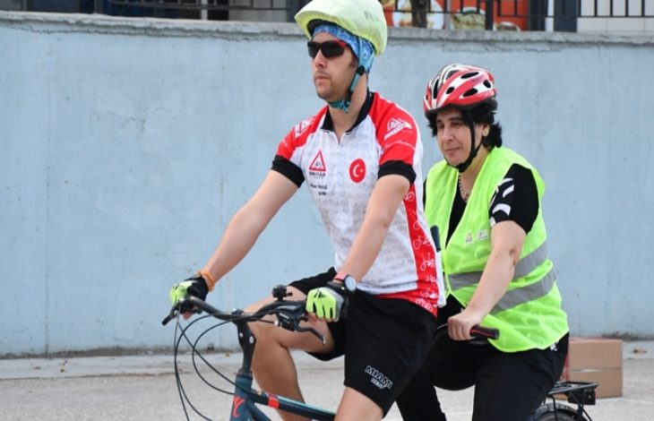 Fotoğrafta iki kişi iki kişilik bir tandem bisiklete biniyor. Ön tarafta kask ve güneş gözlüğü takan bir kişi, arka tarafta ise fosforlu yelek giymiş bir kişi var.