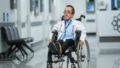 Görselde, beyaz önlük giymiş genç bir doktor adayı tekerlekli sandalyesiyle hastane koridorunda ilerliyor. Boynunda stetoskop ve kimlik kartı bulunuyor. Arkada modern bir hastane ortamı, koltuklar ve danışma bölümü görülüyor.