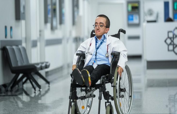 Görselde, beyaz önlük giymiş genç bir doktor adayı tekerlekli sandalyesiyle hastane koridorunda ilerliyor. Boynunda stetoskop ve kimlik kartı bulunuyor. Arkada modern bir hastane ortamı, koltuklar ve danışma bölümü görülüyor.