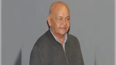 Görselde, yaşlı bir adam gri arka plan önünde ayakta duruyor. Üzerinde koyu renk bir ceket ve kareli gömlek var. Yüzünde hafif bir tebessüm ifadesi bulunuyor.