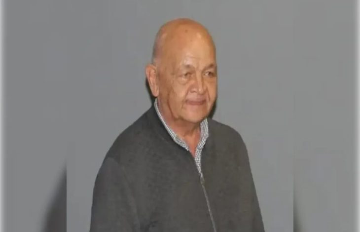 Görselde, yaşlı bir adam gri arka plan önünde ayakta duruyor. Üzerinde koyu renk bir ceket ve kareli gömlek var. Yüzünde hafif bir tebessüm ifadesi bulunuyor.