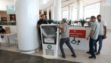 Görselde, bir alışveriş merkezinin içerisinde, **Diyarbakır Büyükşehir Belediyesi** tarafından kurulan **akülü sandalye şarj istasyonu** görülüyor. Üzerinde belediyenin logosu ve bilgilendirme yazısı yer alan cihaz, henüz ambalajı açılmadan yerleştiriliyor. Birkaç görevli erkek, cihazı taşımakta veya yerleştirmekte. Arka planda ise “Engelli Sandalye Şarj Alanı” yazılı bir pano bulunuyor.
