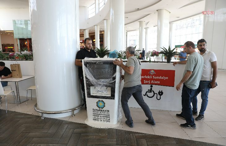 Görselde, bir alışveriş merkezinin içerisinde, **Diyarbakır Büyükşehir Belediyesi** tarafından kurulan **akülü sandalye şarj istasyonu** görülüyor. Üzerinde belediyenin logosu ve bilgilendirme yazısı yer alan cihaz, henüz ambalajı açılmadan yerleştiriliyor. Birkaç görevli erkek, cihazı taşımakta veya yerleştirmekte. Arka planda ise “Engelli Sandalye Şarj Alanı” yazılı bir pano bulunuyor.