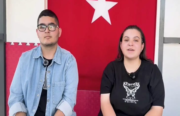 Görselde, arka planda Türk bayrağı bulunan bir ortamda yan yana oturan iki genç görülüyor. Soldaki genç erkek gözlük takıyor, siyah tişört üzerine açık mavi bir ceket giymiş. Sağdaki genç kadın ise siyah tişört giymekte; üzerinde "Kindness" yazısı ve bir kelebek deseni yer alıyor. Her ikisinin de yakalarında yaka mikrofonu var, bu da bir röportaj ya da video kaydı yapıldığını düşündürüyor. İkisi de kameraya dönük bir şekilde sakin ve kendinden emin bir duruş sergiliyor.
