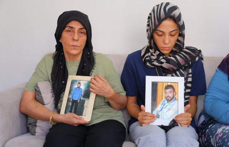 Fotoğrafta iki kadın, ellerinde çerçeveli fotoğraflar tutarak oturuyor; yüzlerinde derin bir üzüntü ifadesi var. Bu kare, yakınını kaybetmiş bir ailenin acısını ve adalet arayışını yansıtıyor.