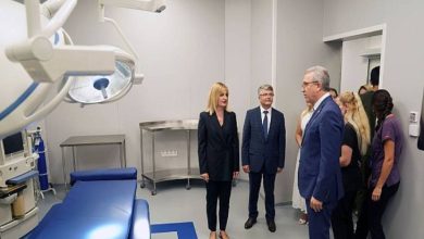 Görselde bir grup kişi modern donanımlı bir hastane odasında inceleme yapıyor. Ön planda ameliyat lambası ve hasta yatağı görülüyor. Takım elbiseli yöneticiler ve sağlık personeli odada ayakta durarak görüşme gerçekleştiriyor. Ortam temiz, düzenli ve profesyonel bir sağlık tesisi izlenimi veriyor.