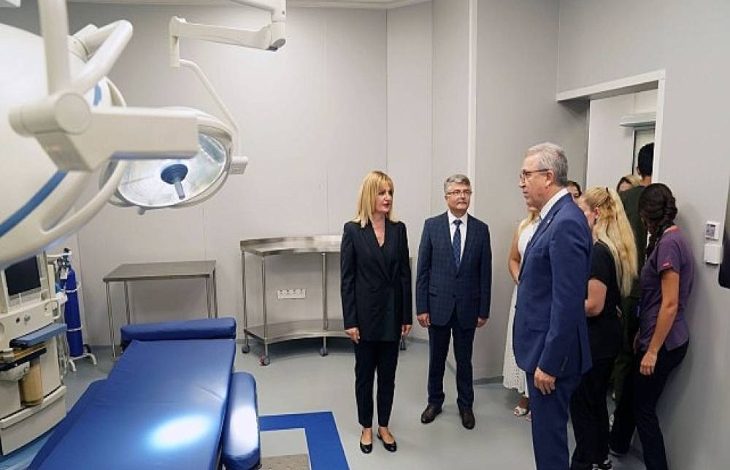 Görselde bir grup kişi modern donanımlı bir hastane odasında inceleme yapıyor. Ön planda ameliyat lambası ve hasta yatağı görülüyor. Takım elbiseli yöneticiler ve sağlık personeli odada ayakta durarak görüşme gerçekleştiriyor. Ortam temiz, düzenli ve profesyonel bir sağlık tesisi izlenimi veriyor.