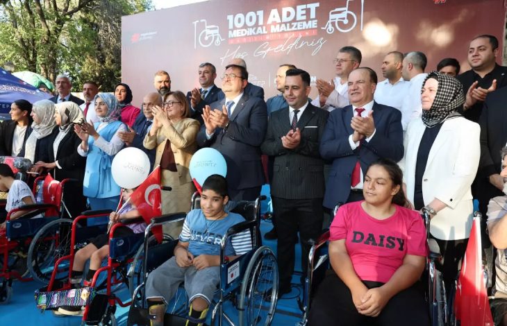 Arka planda "1001 Adet Medikal Malzeme Hoş Geldiniz" yazılı bir afişin önünde, tekerlekli sandalye kullanan çocuklar ve onları destekleyen çok sayıda kamu görevlisi ve vatandaş birlikte poz veriyor. Katılımcılar arasında takım elbiseli erkekler, kadınlar ve çocuklarla birlikte etkinliğe katılan gönüllüler bulunuyor. Topluluk alkış yapıyor.