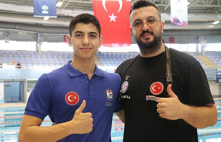 Görselde, bir yüzme havuzu önünde poz veren iki erkek yer alıyor. Soldaki genç sporcu, mavi renkte Türkiye ve federasyon amblemleri bulunan bir polo tişört giymiş, sağ baş parmağını kaldırarak gülümsüyor. Sağdaki kişi ise siyah tişört giymiş, üzerinde Türk bayrağı ve "Spor Toto" logosu yer alıyor. O da baş parmağını kaldırarak poz veriyor. Arka planda ise tribünler, Türk bayrağı ve yüzme havuzu görülüyor.