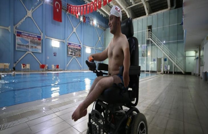 Görselde kapalı bir yüzme havuzunun kenarında tekerlekli sandalyede oturan genç bir sporcu bulunuyor. Başında bone var ve üst kısmı çıplak. Arka planda havuz, merdivenler, Türk bayrakları ve mavi duvarlı bir salon dikkat çekiyor. Sporcu gülümseyerek havuza doğru bakıyor.