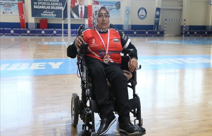 Görselde başörtülü Yeter Olcay, kırmızı ve siyah spor forması giymiş halde tekerlekli sandalyesinde oturuyor. Boynunda madalya asılı. Spor salonunun ortasında duruyor ve arka planda pankartlar ile Türk bayrağı görünüyor.