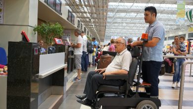 Görselde bir havalimanı terminalinde yolcular işlem sırası bekliyor. Ön planda yaşlı bir yolcu tekerlekli sandalye ile görevli bir personel tarafından bankoya götürülüyor. Arkada ise check-in işlemleri için sırada bekleyen başka yolcular görünüyor. Ortam kalabalık ve hareketli.
