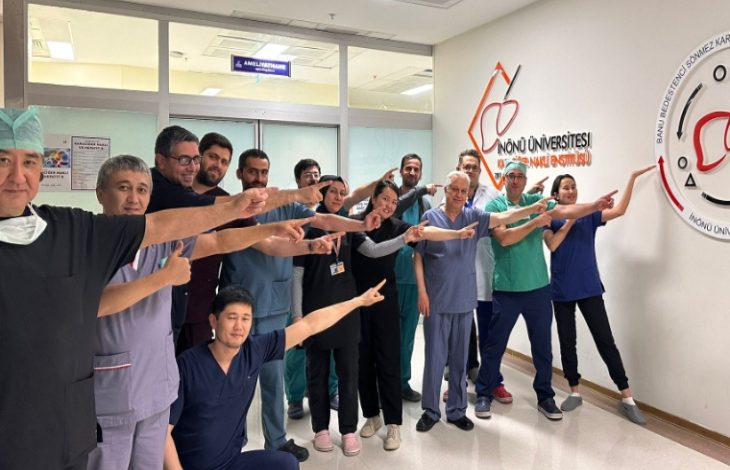 Görselde, ameliyathane kapısının önünde bir grup sağlık çalışanı (doktorlar, hemşireler ve diğer personel) poz veriyor. Hepsi aynı yöne, duvarda yer alan “İnönü Üniversitesi Karaciğer Nakli Enstitüsü” logosuna parmaklarıyla işaret ediyor. Gruptaki herkes mutlu ve gururlu görünüyor.