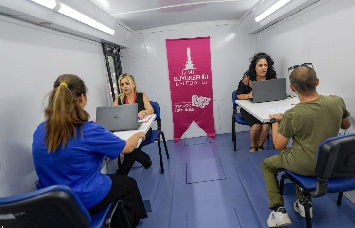 Görselde, İzmir Büyükşehir Belediyesi’ne ait **Meslek Fabrikası Mobil İstihdam Aracı** içinde dört kişi karşılıklı masalarda oturmuş şekilde görülüyor. İki belediye personeli, karşılarındaki bireylerle laptoplar üzerinden görüşme yapıyor. Aracın arka kısmında **İzmir Büyükşehir Belediyesi** ve **Meslek Fabrikası** logolarının yer aldığı pembe bir afiş bulunuyor.