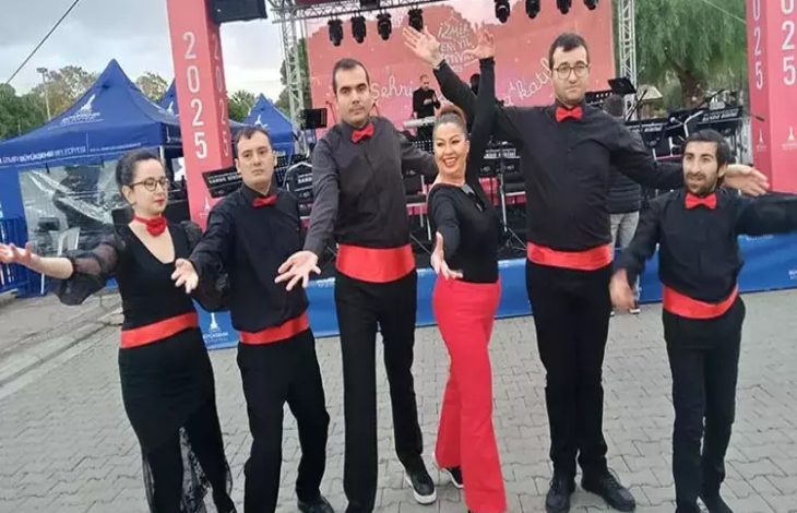 Görselde altı kişilik bir dans ekibi sahne önünde poz veriyor. Hepsi siyah kıyafetler giymiş, kırmızı kuşak ve papyon takmış. Ortam açık hava etkinliğini andırıyor; sahnede müzik ekipmanları ve arka planda etkinlik afişi var.