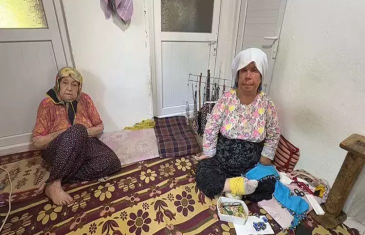 Görselde, sade döşenmiş bir odada yerde oturan iki kadın görülüyor. Solda yaşlı bir kadın, sağda ise başında beyaz örtü olan, engelli olduğu anlaşılan orta yaşlı bir kadın yer almakta. Sağdaki kadın el işi ürünler (boncuk kolyeler, kumaş parçaları) yapıyor gibi görünüyor. Arka planda ise kapılar ve askılığa asılmış takılar yer almakta.