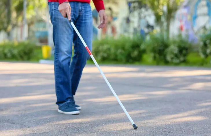 Görselde, açık bir alanda yürüyen görme engelli bir bireyin alt gövdesi görülüyor. Kişi, bir elinde beyaz baston tutarak yolda ilerliyor. Üzerinde kot pantolon ve kırmızı bir üst bulunmakta. Bastonun ucu yere temas etmiş durumda, bu da bireyin yönünü ve yolunu bu baston aracılığıyla belirlediğini gösteriyor. Arka planda yeşillikler ve hafif flu bir çevre manzarası var.