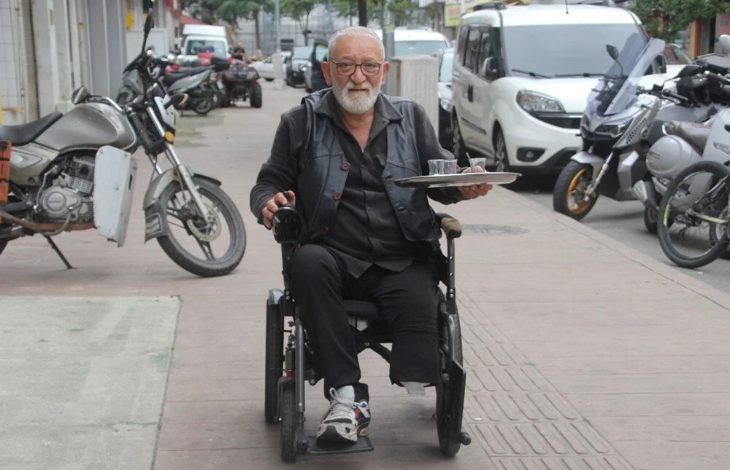 Görselde, akülü tekerlekli sandalye kullanan yaşlı bir adam sokakta ilerlerken görülüyor. Elinde çay bardakları bulunan bir tepsi taşıyor. Arka planda motosikletler, arabalar ve kaldırım üzerindeki yol detayları yer almakta. Adamın yüzünde hafif bir tebessüm var.