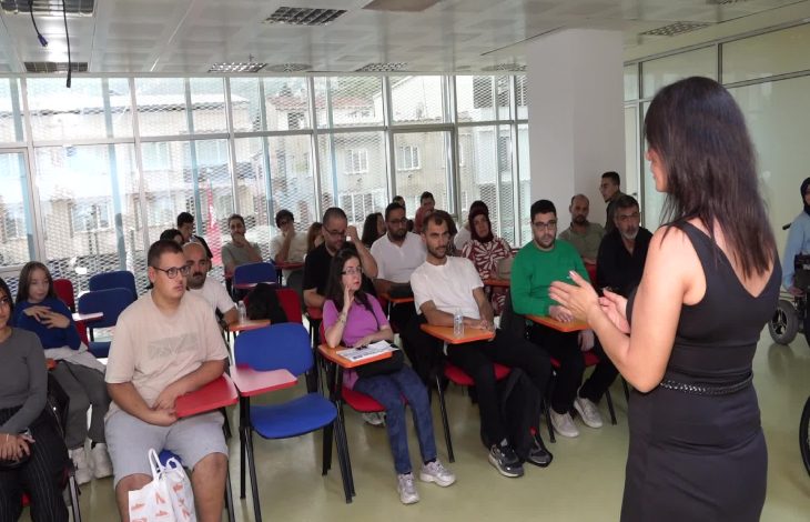Kadın eğitmen kurs salonunda sandalyeler bulunan EKPSS kursiyerlerine ders anlatıyor.
