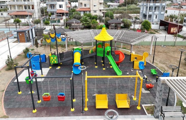 Görselde rengarenk oyun gruplarının yer aldığı modern bir çocuk parkı görülüyor. Parkta kaydıraklar, salıncaklar ve tırmanma alanları bulunuyor. Salıncaklar ve ekipmanlar engelli çocuklar için de uygun şekilde tasarlanmış. Parkın çevresinde binalar ve yeşil alanlar dikkat çekiyor.