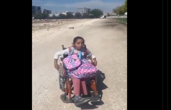 Görselde, okul çantasıyla tekerlekli sandalyede ilerlemeye çalışan küçük bir kız çocuğu, bozuk ve taşlık bir yolda görülüyor. Arka planda yerleşim yerleri bulunmakta. Kare, erişilebilir olmayan yolların engelli çocuklar için nasıl bir zorluk yarattığını çarpıcı şekilde yansıtıyor.