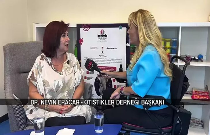 Fotoğrafta, CNN Türk mikrofonuyla röportaj yapan bir muhabir, Otistikler Derneği Başkanı Dr. Nevin Eracar ile bir söyleşi gerçekleştiriyor. Röportaj, bir stüdyo veya danışma odası benzeri, sade ve düzenli bir ortamda gerçekleşiyor. İki kadın karşılıklı oturuyor, masada iki bardak su ve bazı notlar bulunuyor. Arka planda ise “Benimle Oynar Mısın?” adlı bir projeye ait tanıtım afişi dikkat çekiyor.