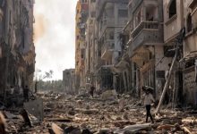 Görselde, savaşın harabeye çevirdiği bir şehir manzarası yer alıyor. Her iki yanda yüksek binalar ağır hasar almış, cepheleri yıkılmış, camlar kırılmış ve enkaz yığınları sokağa saçılmış durumda. Sokakta, yıkıntıların arasında yürümeye çalışan birkaç sivil insan görülüyor; biri başını korur şekilde elini yukarı kaldırmış. Elektrik direkleri devrilmiş, kablolar yerlere sarkmış. Gökyüzü dumanlı, ortam gri ve kasvetli. Bu kare, Gazze’de yaşanan yıkımın boyutunu ve sivil halkın çaresizliğini çarpıcı bir şekilde gözler önüne seriyor.