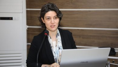 Görselde, Tokat Gaziosmanpaşa Üniversitesi Eğitim Fakültesi Özel Eğitim Bölümü Öğretim Üyesi Dr. Neslihan Can Polat, bir konuşma yaparken görülüyor. Önünde bir dizüstü bilgisayar (Asus marka) ve bir mikrofon bulunmakta. Arka planda ahşap görünümlü bir panel duvar var. Dr. Polat, ciddi ve kararlı bir ifadeyle dinleyicilere hitap ediyor.