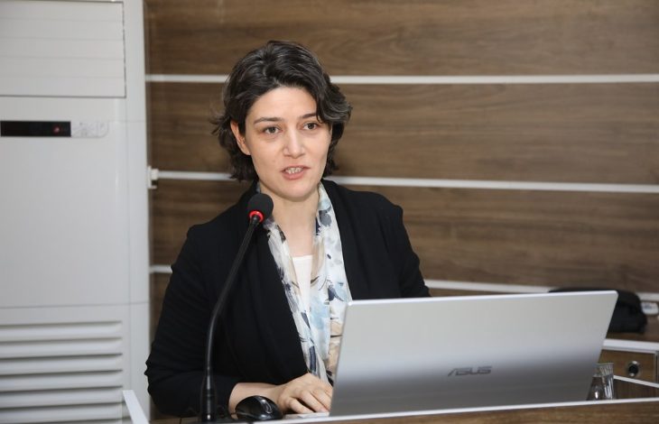 Görselde, Tokat Gaziosmanpaşa Üniversitesi Eğitim Fakültesi Özel Eğitim Bölümü Öğretim Üyesi Dr. Neslihan Can Polat, bir konuşma yaparken görülüyor. Önünde bir dizüstü bilgisayar (Asus marka) ve bir mikrofon bulunmakta. Arka planda ahşap görünümlü bir panel duvar var. Dr. Polat, ciddi ve kararlı bir ifadeyle dinleyicilere hitap ediyor.