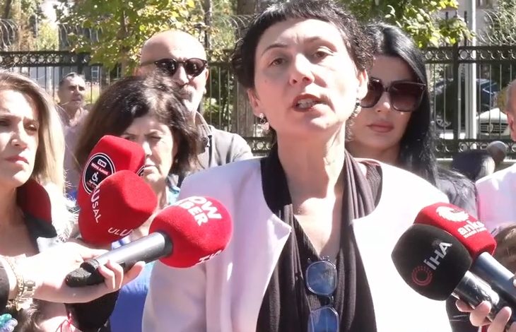 Görselde, bir kadın mikrofonlar önünde basın açıklaması yapıyor. Etrafında toplanmış insanlar bulunuyor. Mikrofonlarda çeşitli haber ajanslarının logoları (İHA, Show Haber, Ulusal Kanal) görülüyor. Açıklama, açık havada ve kalabalık bir grup önünde gerçekleştiriliyor.