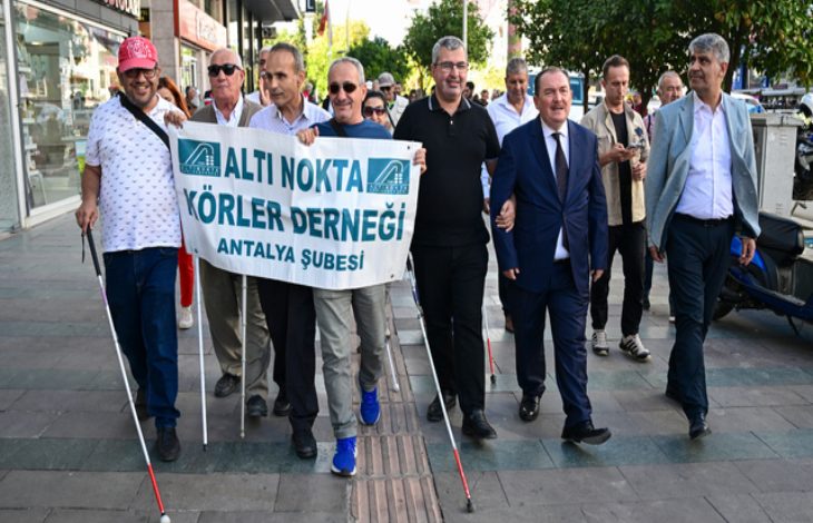 Görselde bir grup kişi kaldırımda yürüyüş yapıyor. Ön sırada “Altı Nokta Körler Derneği Antalya Şubesi” yazılı bir pankart taşıyan kişiler var. Bazı katılımcılar beyaz baston kullanıyor. Arka planda diğer yürüyüşçüler ve şehir ortamı görülüyor. Katılımcılar gülümseyerek birlikte ilerliyor.