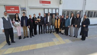Görselde, bir grup kişi bina önünde hatıra fotoğrafı çekiliyor. Aralarında kadınlar ve erkekler bulunuyor, bazı kişiler çiçek buketi tutuyor. Arkadaki duvarda “Camii Engelsiz Girişi” ve “Engelsiz Kur’an Kursu” yazılı tabelalar görülüyor.