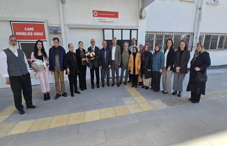 Görselde, bir grup kişi bina önünde hatıra fotoğrafı çekiliyor. Aralarında kadınlar ve erkekler bulunuyor, bazı kişiler çiçek buketi tutuyor. Arkadaki duvarda “Camii Engelsiz Girişi” ve “Engelsiz Kur’an Kursu” yazılı tabelalar görülüyor.