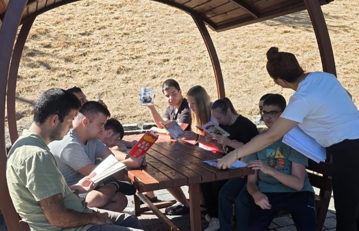 Bir grup genç, ahşap bir kamelya altında oturarak kitap okuyor. Bir kadın onlara rehberlik ediyor ve elindeki belgelerden bir şeyler gösteriyor. Arka planda kuru otlarla kaplı bir yamaç görülüyor.