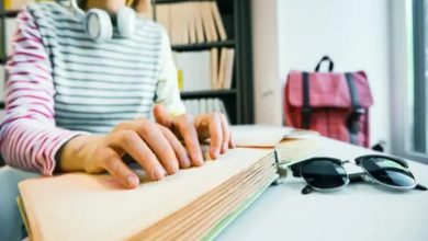 Görselde, bir kişi Braille alfabesiyle yazılmış bir kitabı elleriyle okuyor. Üzerinde kulaklık, çizgili bir tişört var. Masada bir güneş gözlüğü duruyor, arka planda raflar ve pembe bir çanta görülüyor.