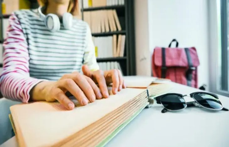 Görselde, bir kişi Braille alfabesiyle yazılmış bir kitabı elleriyle okuyor. Üzerinde kulaklık, çizgili bir tişört var. Masada bir güneş gözlüğü duruyor, arka planda raflar ve pembe bir çanta görülüyor.