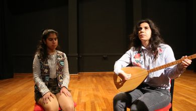 Görselde sahne üzerinde iki genç kadın oturuyor. Sağda oturan kadın bağlama çalıyor, soldaki ise gözleri kapalı şekilde dinliyor. Her ikisi de sahne ışığı altında kırmızı sandalyelerde oturmakta. Arka plan siyah, zemin ise ahşap kaplamalı. Ortam bir müzik performansı ya da prova alanını andırıyor.