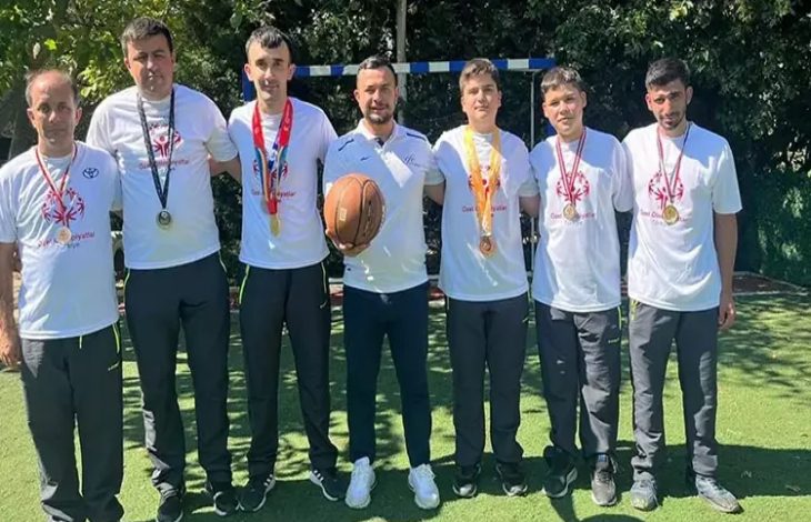 Görselde, açık havada bir araya gelmiş bir grup sporcu ve antrenör görülüyor. Üzerlerinde beyaz tişörtler bulunan sporcuların boyunlarında kazandıkları madalyalar asılı. Ortadaki kişi elinde bir basketbol topu tutuyor. Arkalarında ise küçük bir kale direği ve ağaçlar yer almakta. Sporcuların mutlu ve gururlu olduğu dikkat çekiyor.