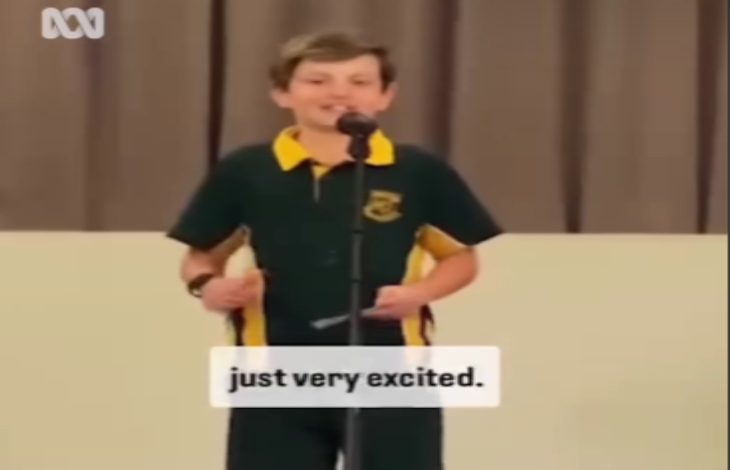 Görselde, okul forması giymiş bir çocuk bir sahnede mikrofonun önünde konuşma yaparken görülüyor. Alt kısımda “just very excited.” yazılı altyazı yer alıyor. Arka planda kahverengi bir perde bulunuyor.