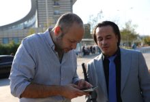 Görselde, açık havada iki erkek yan yana duruyor. Soldaki kişi cep telefonuna bakarken, sağdaki kişi gri takım elbise ve mavi kravat giymiş şekilde ona eşlik ediyor. Arka planda modern bir bina ve birkaç kişi görünüyor.