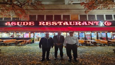 Görselde akşam saatlerinde bir restoranın önünde duran üç erkek görülüyor. Restoranın tabelasında kırmızı renkle “ASUDE RESTAURANT” yazıyor. Etrafta sonbahar yaprakları yerde birikmiş ve dış mekanda ahşap masalar bulunuyor. Arka planda restoranın içi ışıklarla aydınlatılmış şekilde görünüyor.