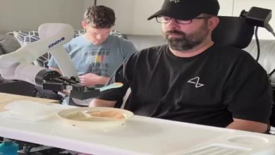 Bir erkek, tekerlekli sandalyede otururken önündeki masada bulunan robotik bir kol yardımıyla yemek yiyor. Robot kol, kaşığı tutarak tabaktaki yemeği kişiye doğru uzatıyor. Arka planda başka bir kişi telefonuyla ilgileniyor.