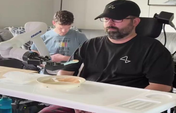 Bir erkek, tekerlekli sandalyede otururken önündeki masada bulunan robotik bir kol yardımıyla yemek yiyor. Robot kol, kaşığı tutarak tabaktaki yemeği kişiye doğru uzatıyor. Arka planda başka bir kişi telefonuyla ilgileniyor.