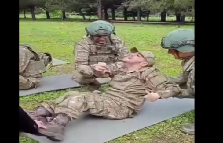 Askerî kıyafetli bir grup asker açık alanda egzersiz yapıyor. Ortada yerde sırtüstü yatan bir asker mekik çekerken, yanındaki diğer askerler ona moral veriyor ve destek oluyor.