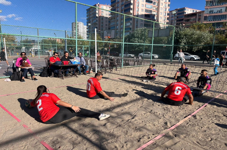 Görselde açık havada bir oturarak voleybol maçı oynanıyor. Kırmızı formalı ve siyah formalı iki takım oyuncuları sahada oturarak top oynuyor. Arkada hakem masası ve seyirciler, çevrede ise apartmanlar ve yeşillikler görülüyor. Hava güneşli ve açık.