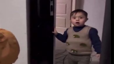 Görselde küçük bir çocuk kapıdan içeri bakıyor. Üzerinde desenli bir süveter ve koyu renk bir kazak var. Yüz ifadesi şaşkınlık ve sevinç karışımı bir duygu yansıtıyor. Arka planda açık bir kapı ve iç mekân detayları görülüyor.