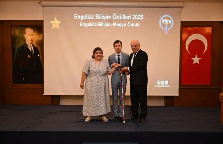 Görselde, “Engelsiz Bilişim Ödülleri 2025” töreninde sahnede üç kişi yer alıyor. Ortadaki kişi ödül alırken diğer iki kişi yanında duruyor. Arkada “Engelsiz Bilişim Medya Ödülü” yazılı bir perde, sol tarafta Atatürk portresi ve sağda Türk bayrağı bulunuyor.