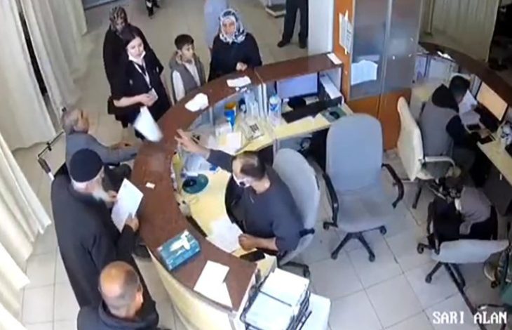 Görselde, yarım daire şeklinde bir danışma masası etrafında çeşitli insanlar toplanmış. Masanın arkasında görevli bir doktor oturuyor ve maske takıyor; elinde bir kağıt tutarken karşısındaki kişilere uzatıyor. Masanın önünde bekleyen kişiler arasında yaşlı bir adam, bir çocuk, kadınlar ve birkaç yetişkin erkek bulunuyor.