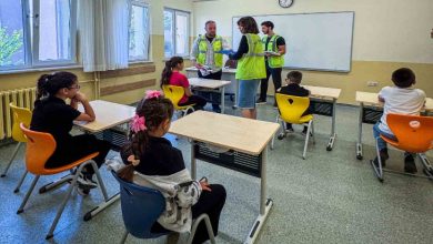 Görselde bir sınıf ortamı görülüyor. Sıralarda oturan çocuklar önde, tahtanın önünde ise yeşil yelek giymiş yetişkinler ayakta duruyor ve öğrencilerle ilgileniyor. Çocuklar dikkatle onları dinlerken, yetişkinler belgeler üzerinde çalışıyor gibi görünüyor. Ortam, eğitim veya bilgilendirme amaçlı bir etkinliği andırıyor.